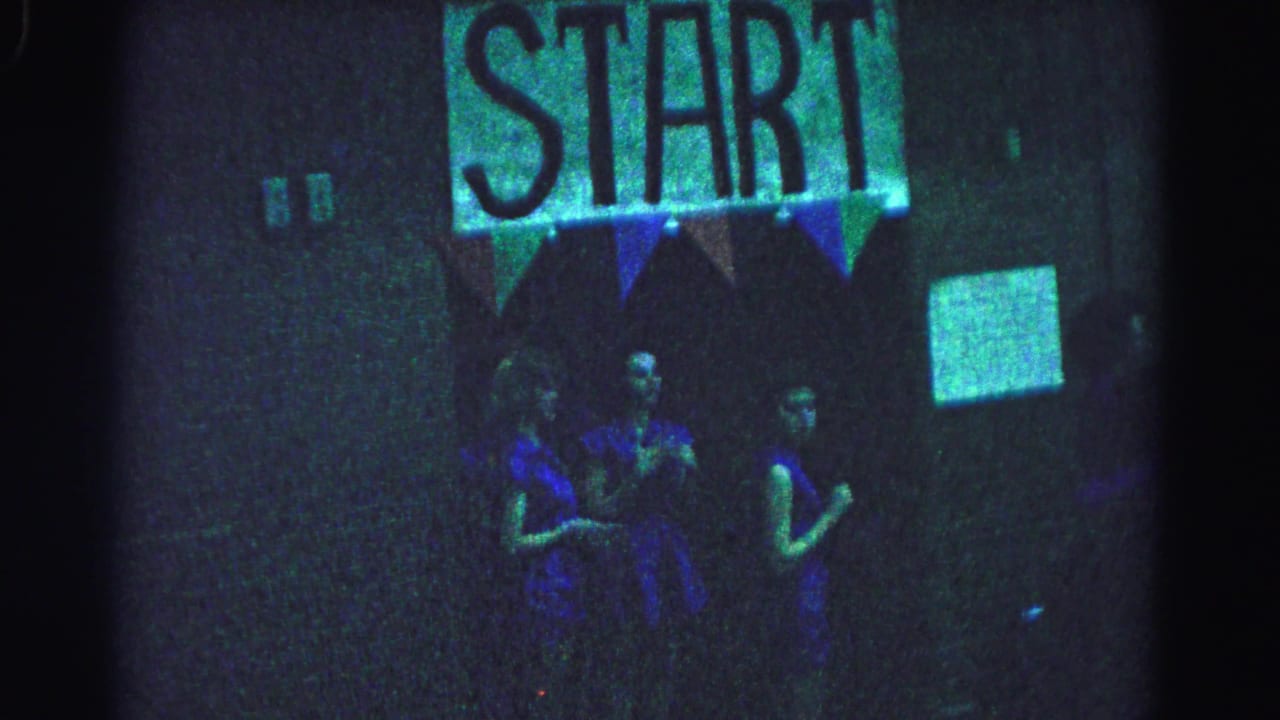 Two teenage girls in blue dresses stand beneath a lit START sign with triangular bunting in a dimly lit indoor space.