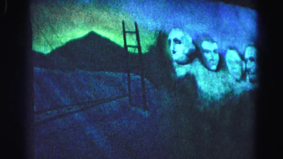 A projected slide of Mount Rushmore appears during a 1963 patriotic event in Hagerstown, Maryland; ladder and blue-green ambient light visible in the darkened venue.