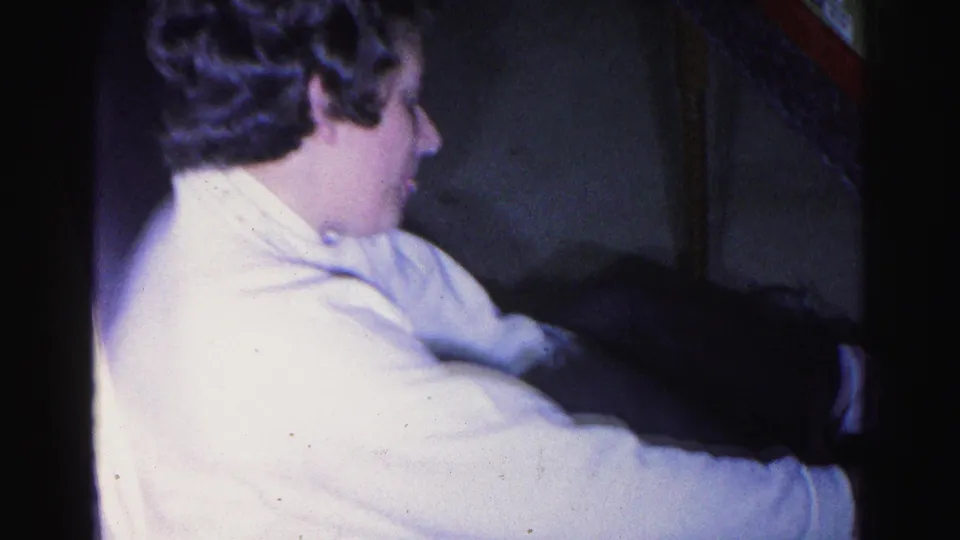 Young woman in profile in white long-sleeved shirt, reaching toward a dark surface in a dim interior; consistent with 1963 8mm home movie
