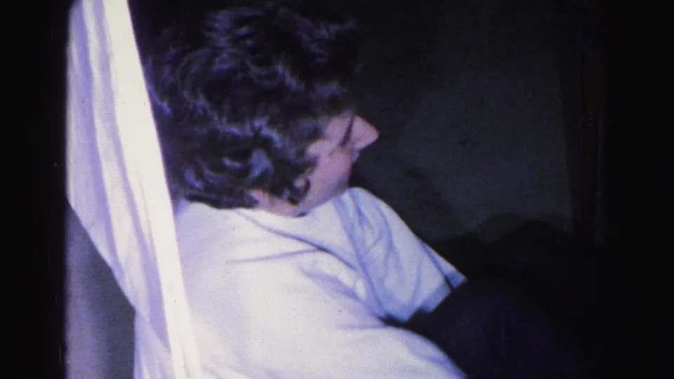 Young woman with dark curly hair and head bowed in white long-sleeved shirt beside white fabric; dim interior, 1963 8mm home movie