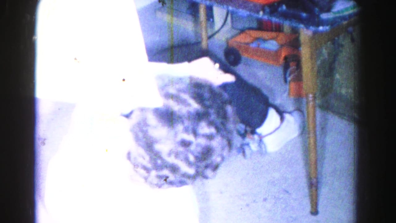Overhead 8mm shot of a young woman with dark curly hair bending forward on a floor, wooden furniture leg and orange household object visible nearby.