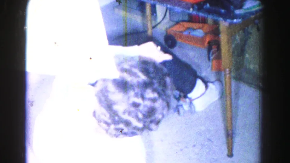 High-angle 8mm home movie frame, c.1963, showing a young woman in a white sweater leaning forward on an interior floor, wooden chair leg and orange object nearby.
