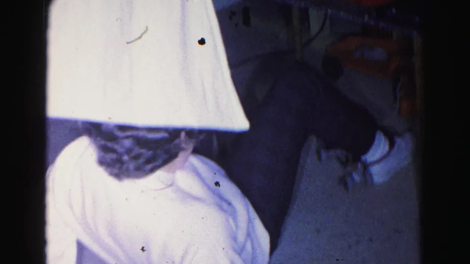 8mm home movie frame c.1963, young woman in white sweater with head bowed, white lampshade dominating the foreground, orange household object at upper right.