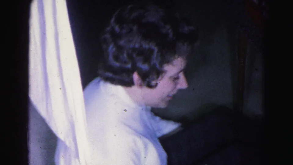 8mm home movie frame c.1963, medium close-up of a young woman in profile wearing a white sweater, dim domestic interior with partial lampshade visible at frame left.