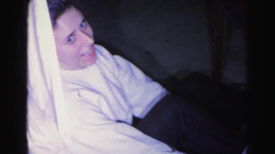8mm home movie frame c.1963, young woman in white sweater looking directly up at the camera with a candid expression, lampshade at left, dark domestic interior behind her.