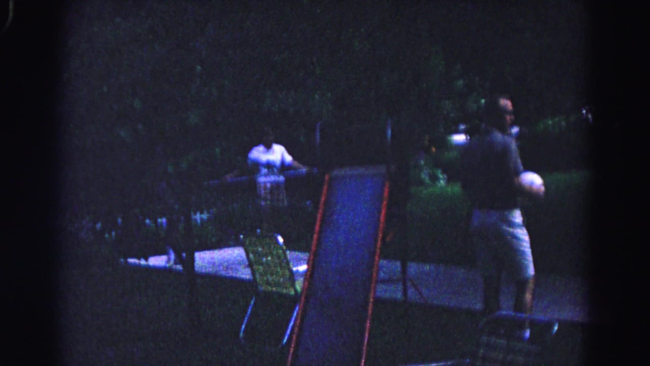 Man in dark shirt holding white ball stands in nighttime backyard beside a small children's slide; second figure visible in background.