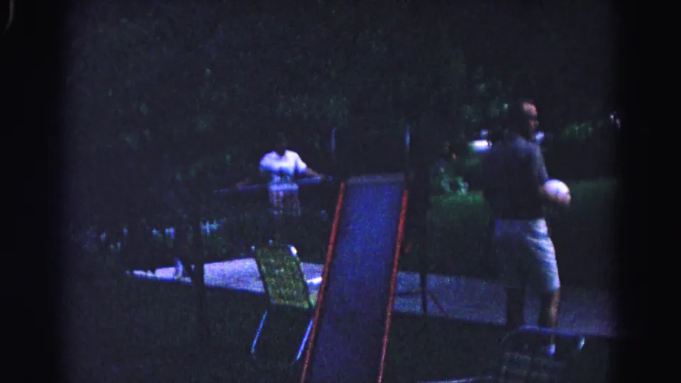 Man holding white ball stands beside a small children's slide in a nighttime backyard, likely Hagerstown, Maryland, circa 1963; folding lawn chair and second figure behind him.