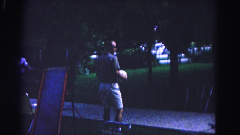 Man holding white ball in a dimly lit nighttime backyard beside a partially visible children's slide, Hagerstown, Maryland, circa 1963; point of artificial light visible in background.