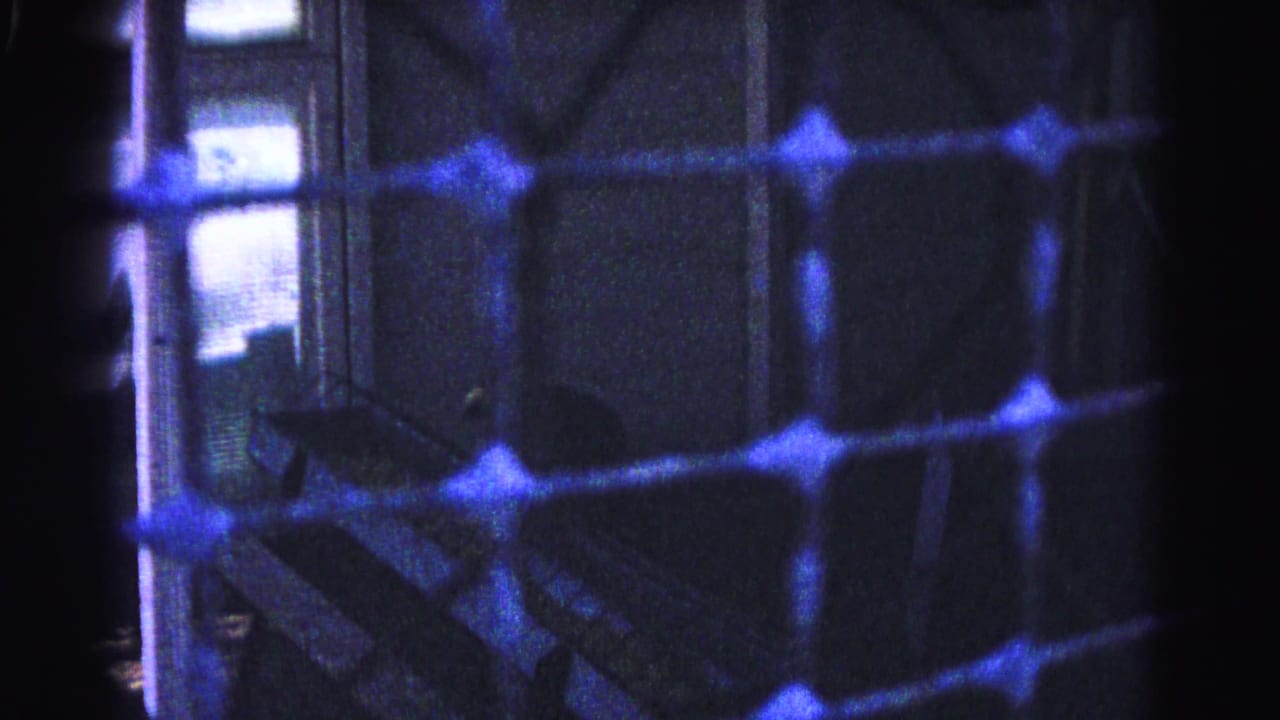 Severely underexposed 8mm frame showing a wire mesh or lattice foreground with vivid blue prismatic flares and a shadowed interior staircase beyond.