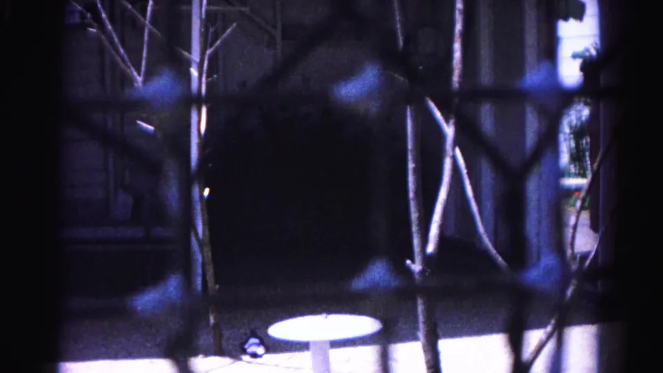 8mm still c.1963, Hagerstown — fence foreground, dark billowing shapes at lower frame, round pale ground object, and residential building at right through blue haze.