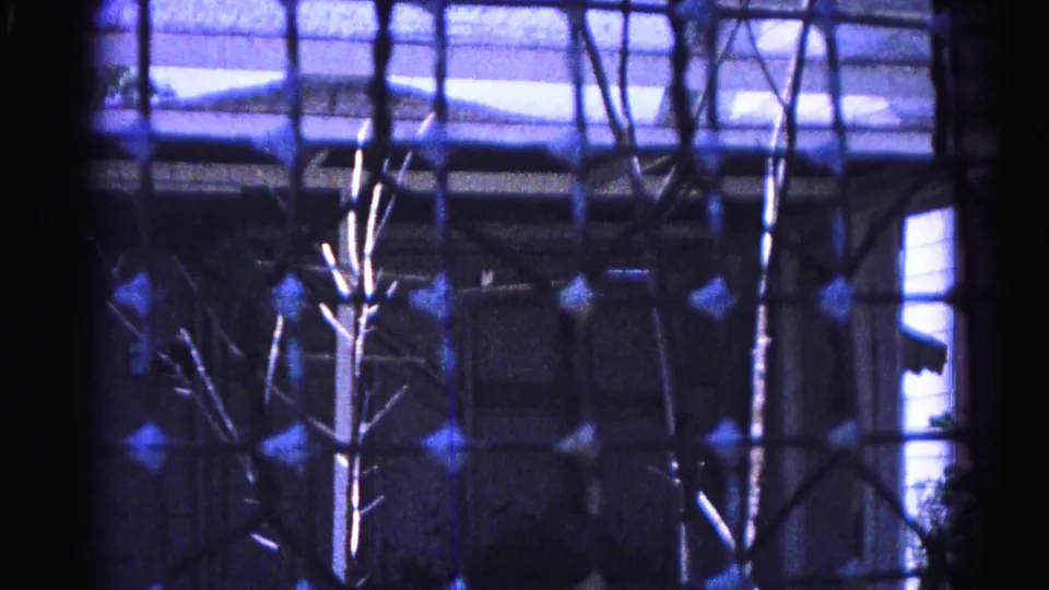 Mid-clip 8mm frame from 1963 Hagerstown home movie, wire screen mesh, leafless branches, and a white round table in the suburban backyard