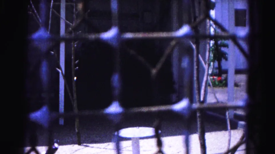 8mm home movie frame circa 1963, Hagerstown Maryland: bare winter branches and chain-link fence foreground a concrete backyard with a small pedestal table and house facade.