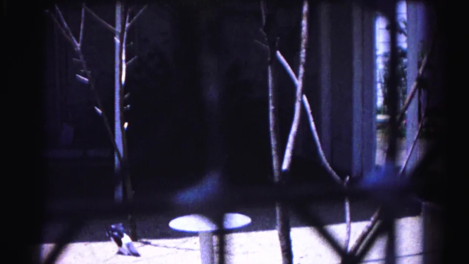 8mm home movie frame circa 1963, Hagerstown Maryland: chain-link fence and leafless branches partially obscure a concrete backyard; small patio table and house structure visible behind.