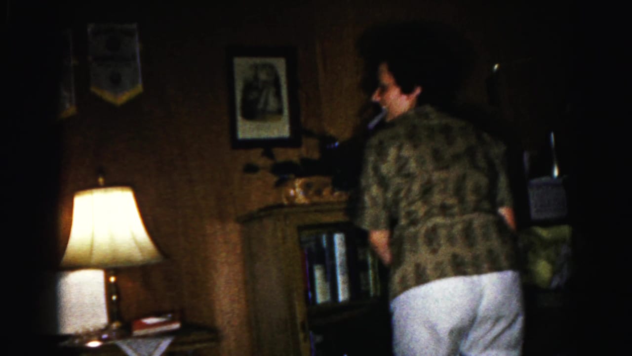 Woman in patterned shirt and white shorts stands with back to camera in a dim wood-paneled living room; lit table lamp visible at left.