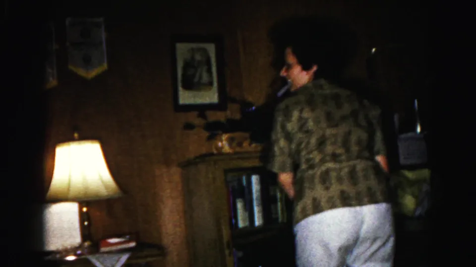 Woman in patterned short-sleeve shirt and white shorts in a wood-paneled living room; lit table lamp and framed pictures visible; 1963 8mm home movie, Hagerstown, Maryland.