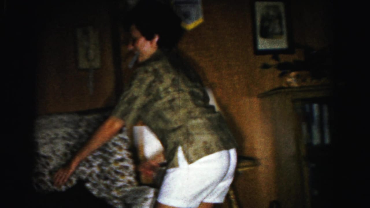 Woman in patterned shirt turns partially toward camera, arms extended, holding a light-colored object in a dim living room.