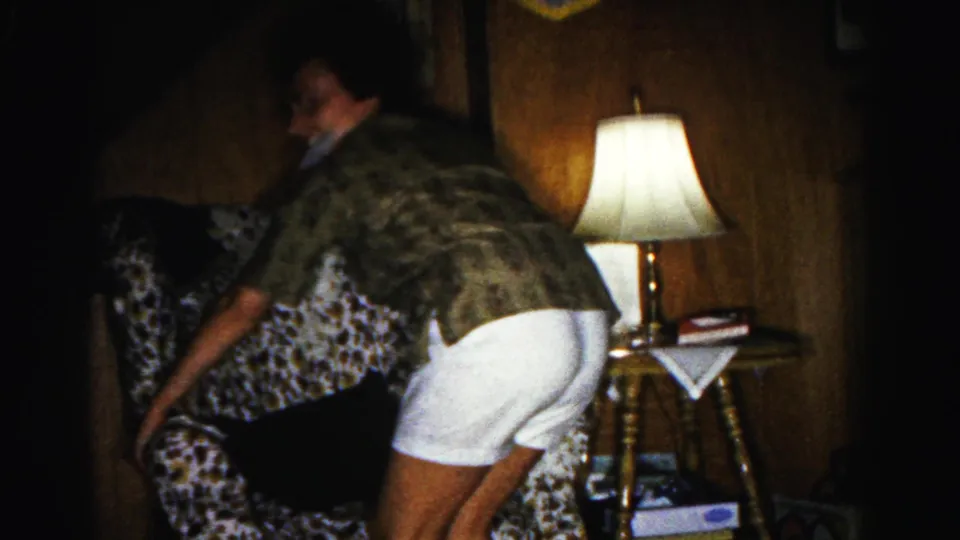 Woman bends to place a dark cushion on a floral-upholstered armchair in a wood-paneled living room; tripod lamp and side table with books visible; 1963 8mm home movie, Hagerstown, Maryland.