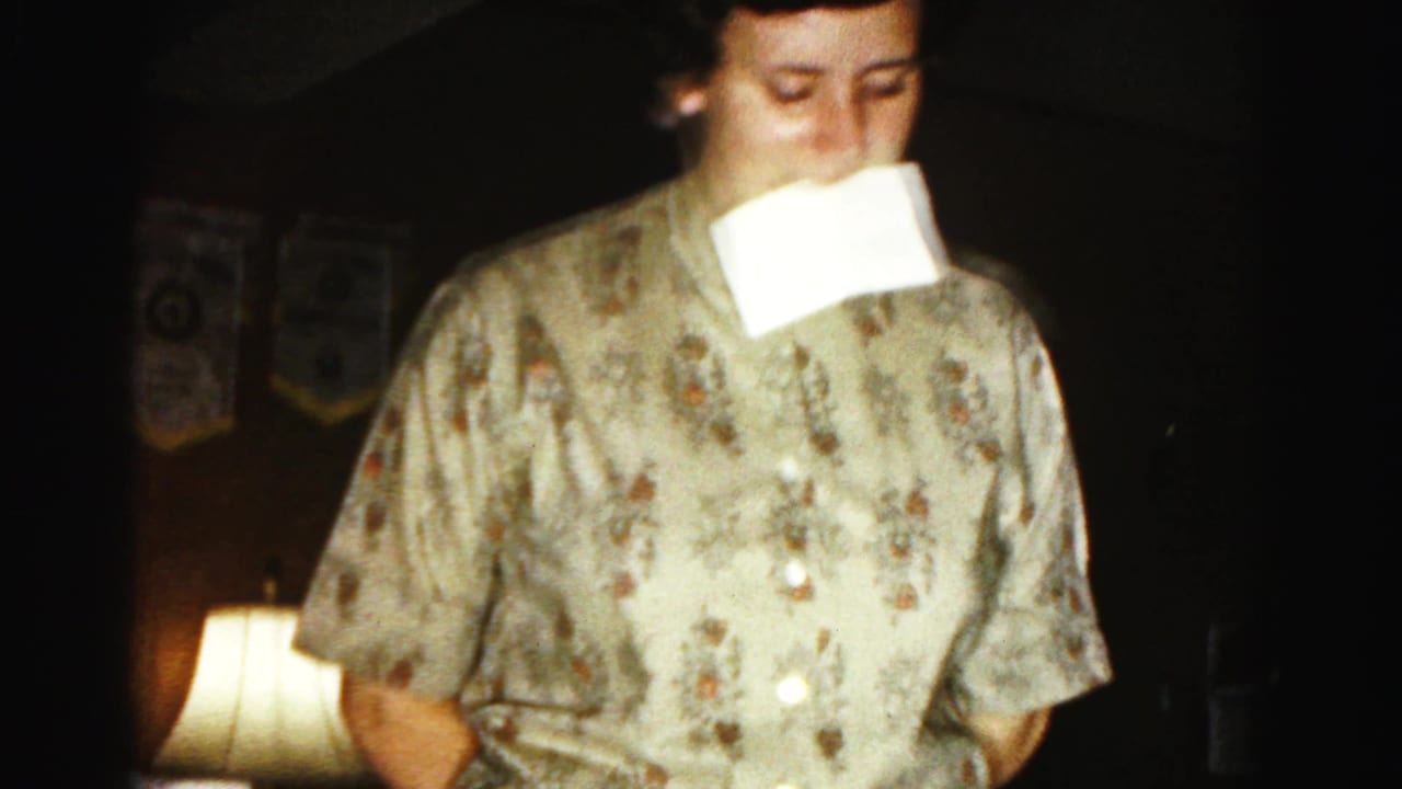 Woman in floral blouse holding white paper in her mouth, standing in a dimly lit living room with a glowing table lamp.
