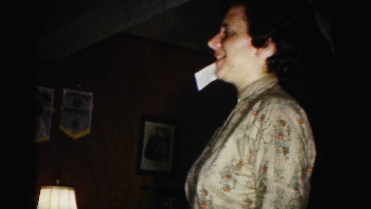 Woman in profile laughing, paper at mouth, blue and yellow pennants and framed items visible on dark-paneled wall behind her.