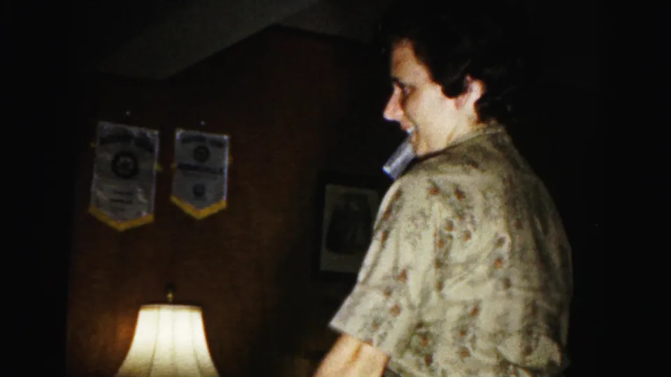 Woman mid-turn smiling broadly during a parlor game in a dimly lit 1960s American home; pennants and table lamp visible on dark-paneled wall.