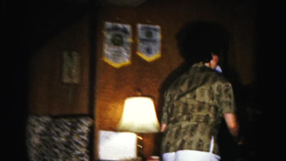 Woman walking away from camera holding paper in a dimly lit 1960s American living room; pennants, glowing table lamp, and upholstered chair visible.