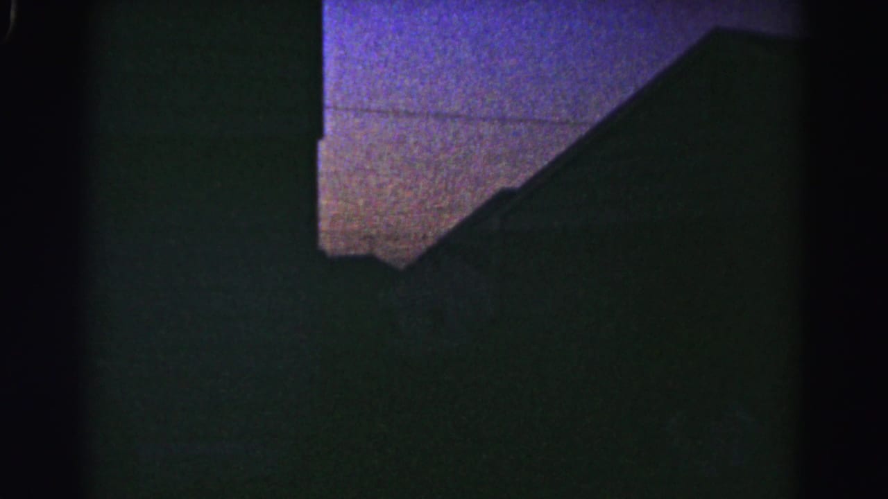 Dark, heavily underexposed 8mm frame showing a dim interior with purple-blue tinted ceiling area and deep shadow throughout.