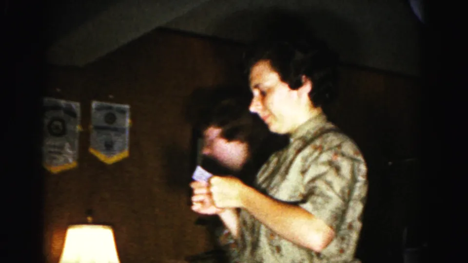 Person acting during a charades game holds a small card before wood-paneled walls with two organizational banners — 1963 home movie, Hagerstown, Maryland.