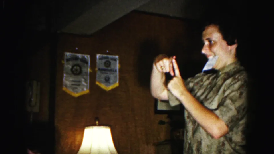 Person mid-gesture during charades, hands at face, smiling — wood-paneled room with two organizational banners visible, 1963 home movie, Hagerstown, Maryland.