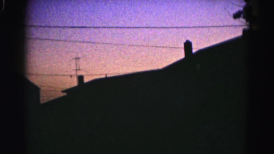 1963 residential skyline view with antennas, chimneys, roof silhouettes, and utility wires beneath pink-blue evening light.