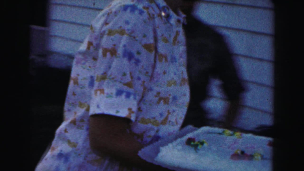 Person in a patterned shirt holds a decorated cake near house siding.