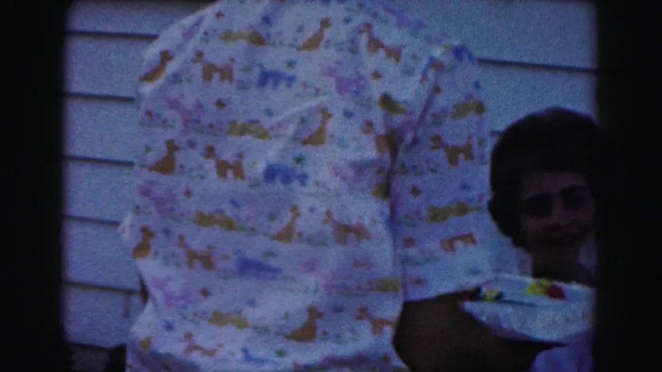1963 Hagerstown home-movie frame showing a cake being carried past a seated guest during a home birthday gathering.