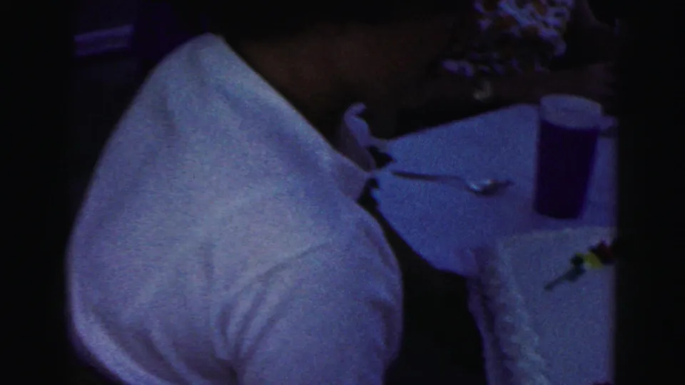 1963 Hagerstown home-movie frame showing a seated party guest at a table as the birthday cake is presented nearby.