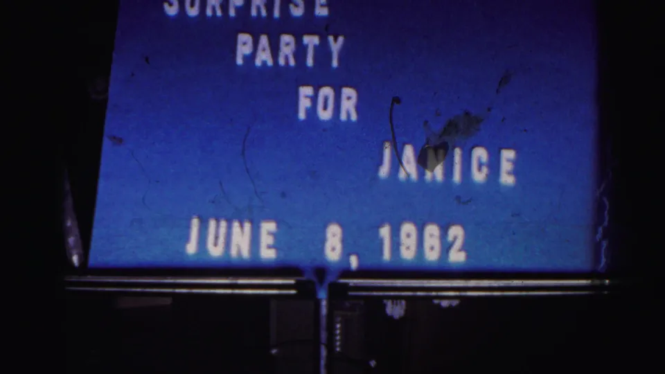 Damaged-looking 8mm frame of the surprise party title card, with dust marks over the blue field and Janice still readable.