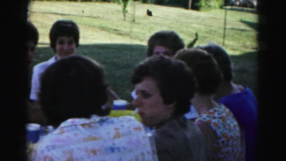 Hagerstown home-movie frame of adults at an outdoor party table in a grassy yard, with 1960s hairstyles and casual clothing visible.