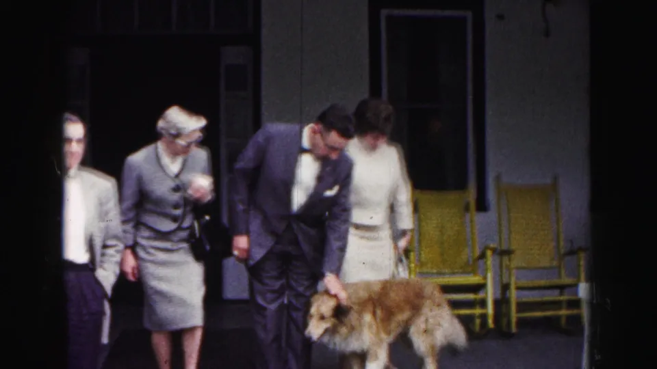 In the Hagerstown 1963 clip, a formally dressed man bends toward a brown dog while other adults stand near yellow porch chairs.