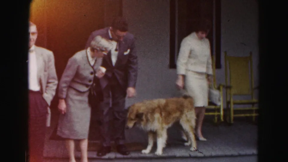 A man in formal clothing rubs a brown dog's head on a porch in 1963 Hagerstown footage, with adults and yellow chairs nearby.