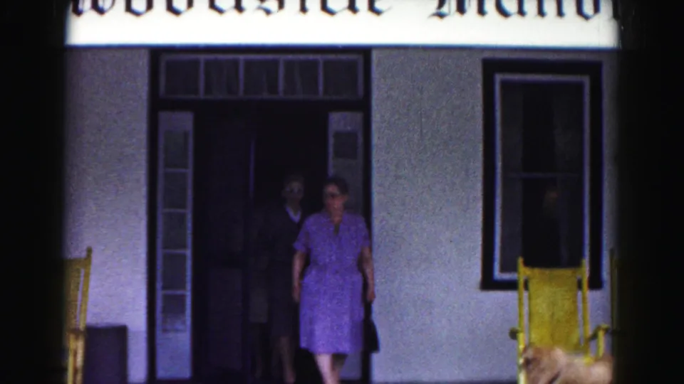 1963 Hagerstown home-movie frame showing a woman in a purple dress stepping out of Woodside Manor with yellow porch chairs nearby.