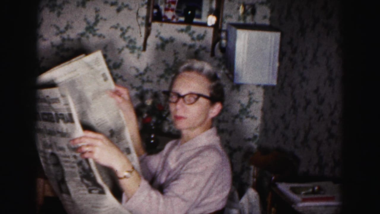 Adult woman in glasses reads a newspaper in a 1963 Hagerstown home interior with floral wallpaper and household furnishings.