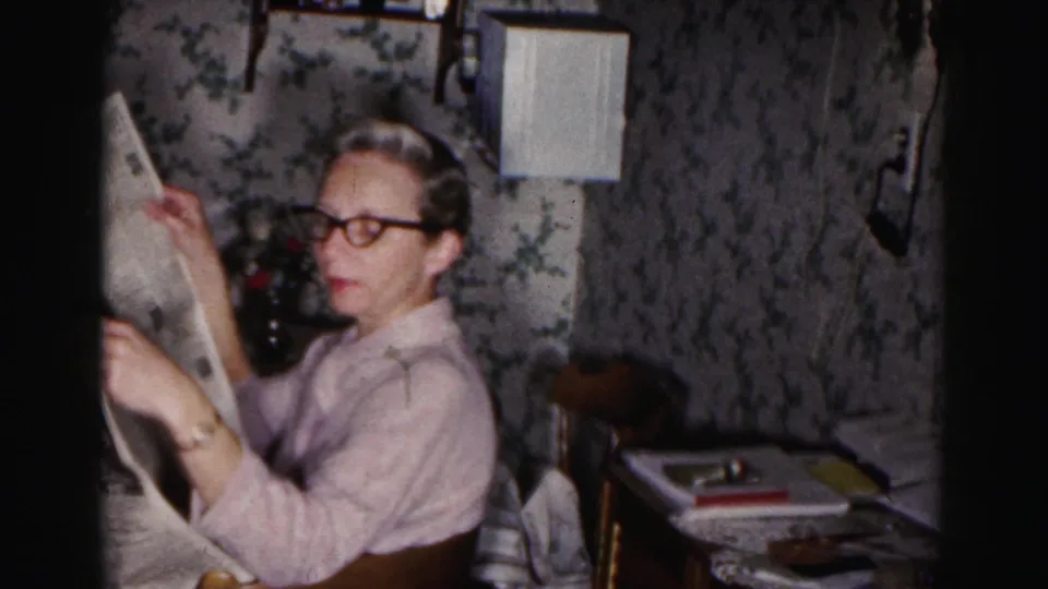 The seated woman continues reading a newspaper in a private 1963 Hagerstown interior with a lamp, wallpaper, and small table nearby.