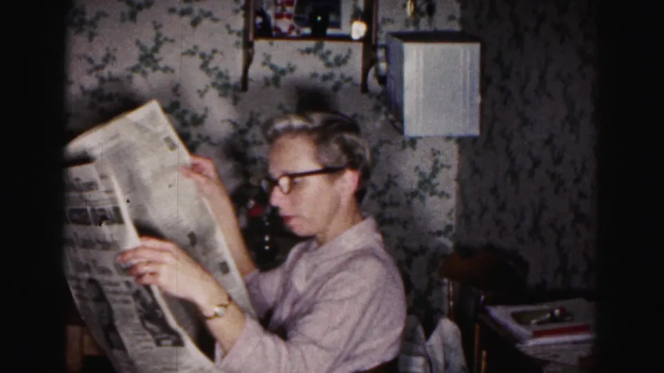 A 1963 home-movie frame shows the woman reading quietly, with dark glasses, floral wallpaper, a wall lamp, and a small table visible.