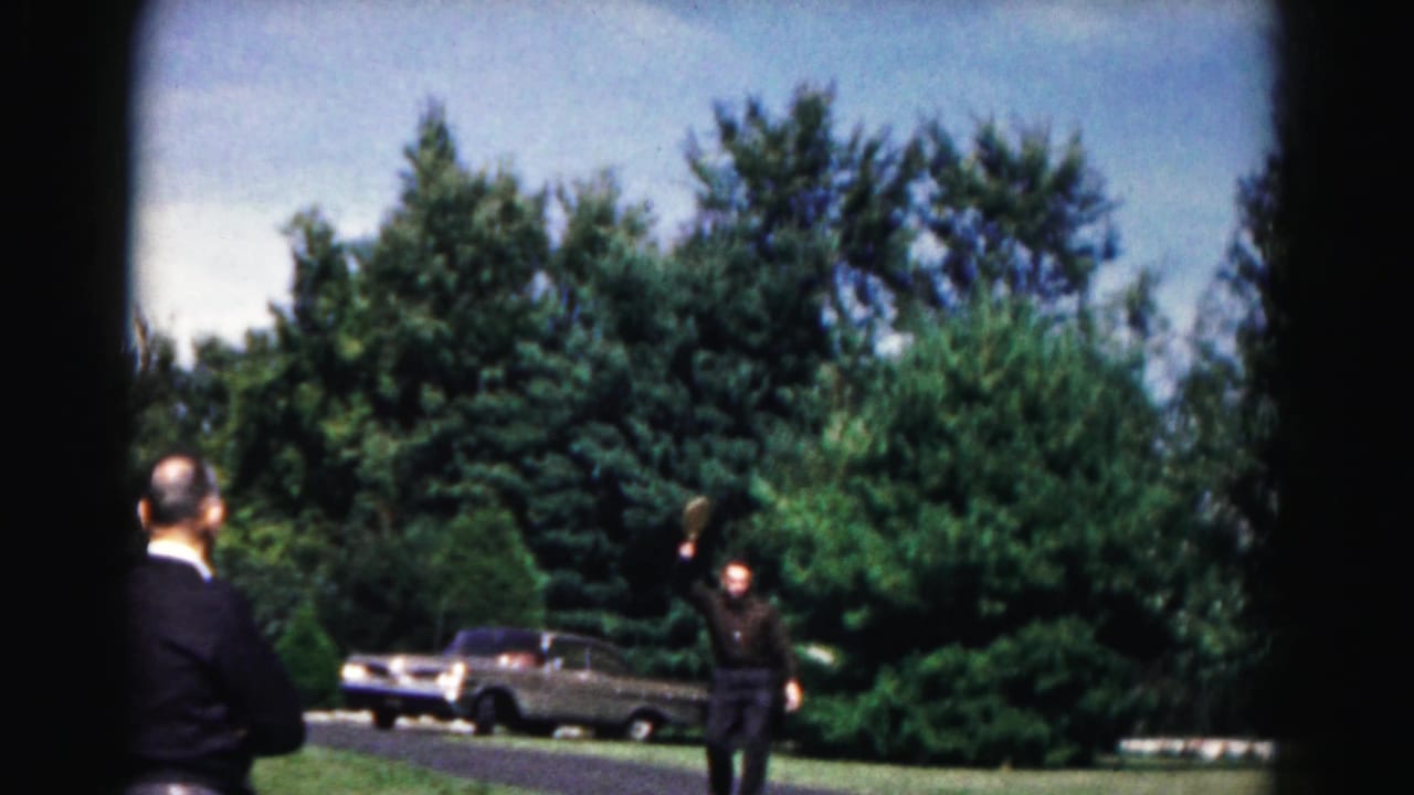 Two men outdoors near a parked vintage car, with one raising an arm on grass beside trees.