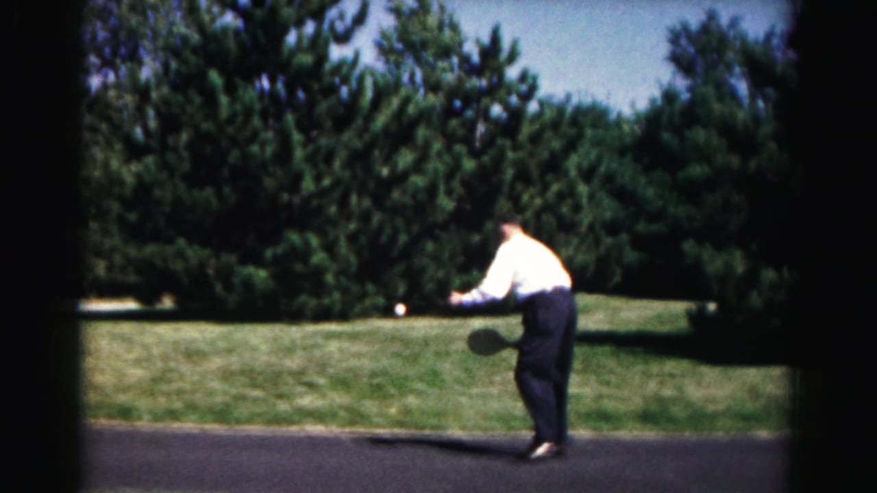 Adult man in a white shirt and dark pants plays paddleball outdoors in Hagerstown, Maryland, with trees and grass behind him.