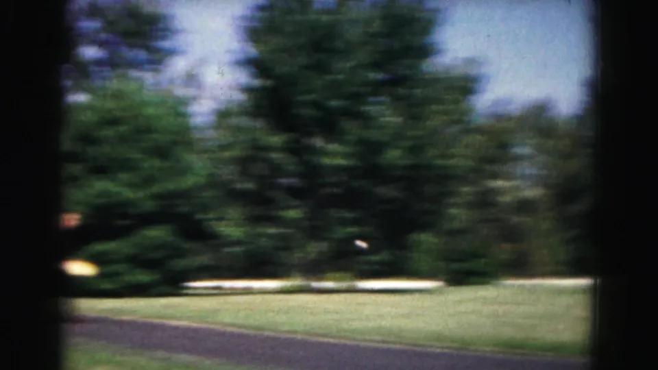 Motion-blurred 8mm frame from the Hagerstown paddleball scene, showing trees, grass, and paved ground as the camera moves quickly.