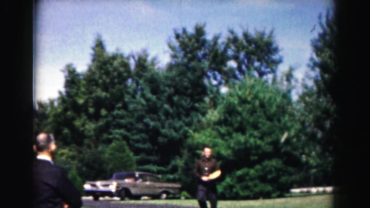 In Hagerstown, Maryland in 1963, a man with a wooden paddle stands on a paved path as another adult watches beside a lawn and parked car.