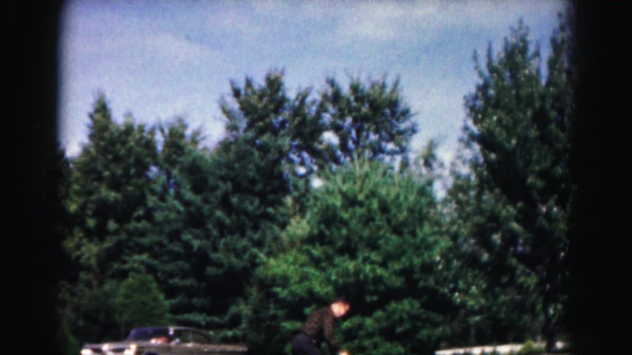 In the 1963 Maryland home-movie scene, the man bends low on the paved path during an outdoor paddle-and-ball game near trees and a parked car.