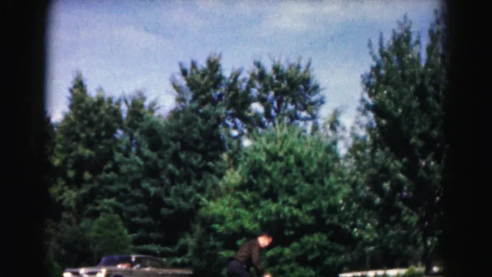 In the 1963 Maryland home-movie scene, the man bends low on the paved path during an outdoor paddle-and-ball game near trees and a parked car.