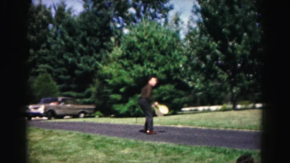 A man in dark clothing plays or practices with a wooden paddle on a paved path in the Hagerstown, Maryland yard scene from 1963.