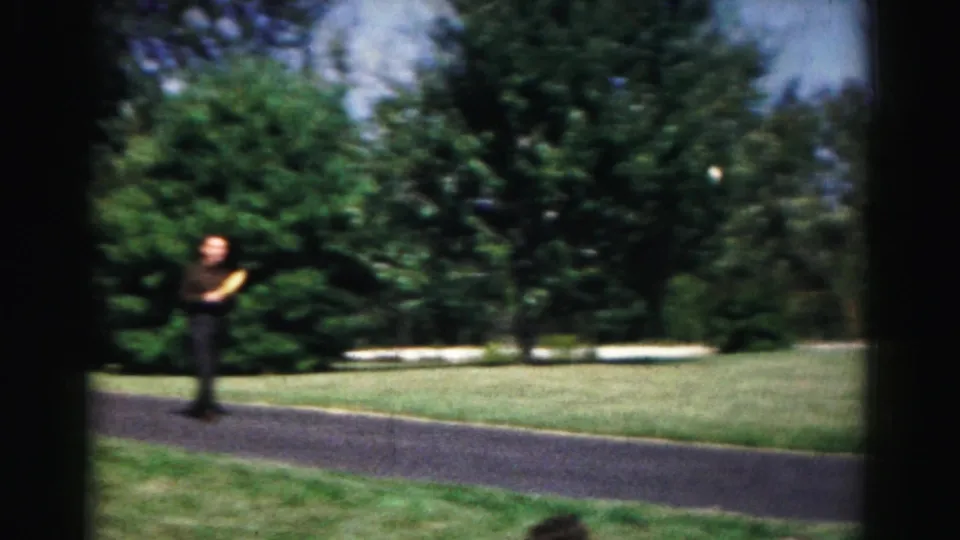 The 1963 Hagerstown home-movie footage shows the man holding a paddle on a paved path bordered by lawn, trees, and shrubs.