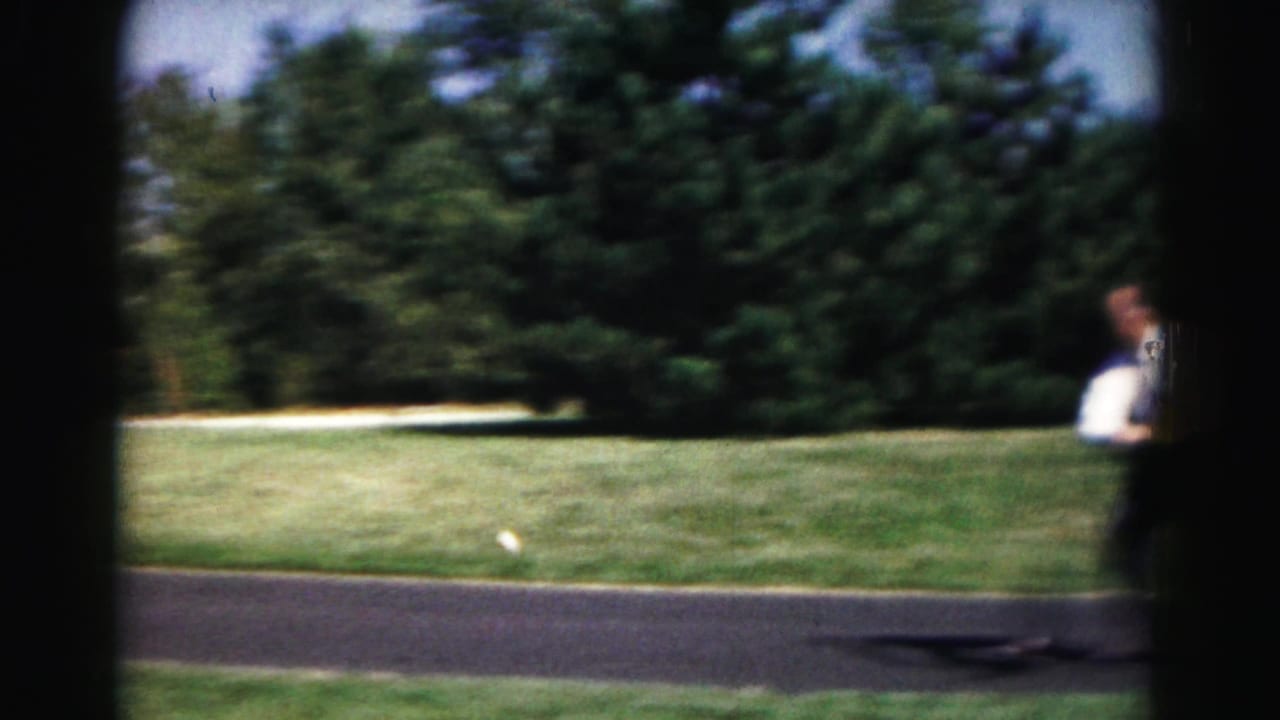 Man in a white dress shirt stands near a paved park path beside grass and trees.
