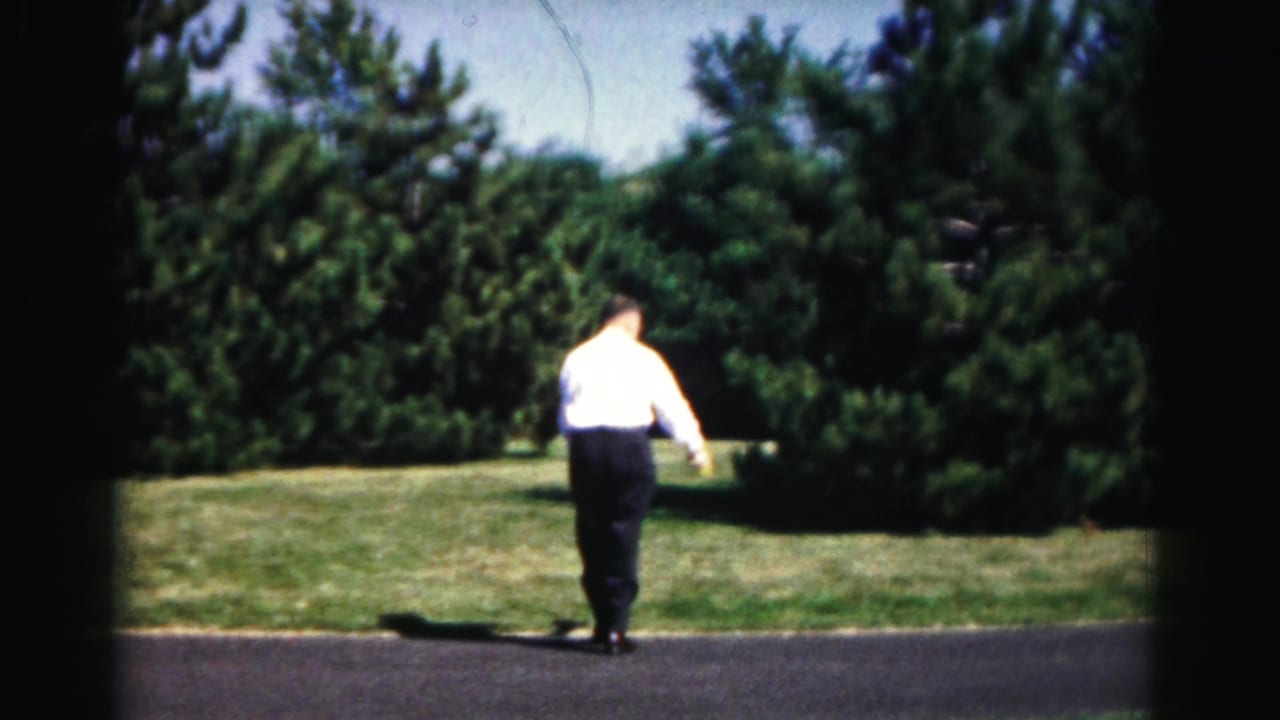 In 1963 Hagerstown, Maryland home-movie footage, a man in a white shirt stands near a paved path with lawns and trees behind him.